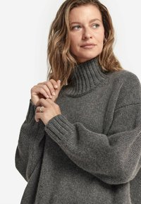 Grauer gestrickter Pullover mit hohem geripptem Kragen und weiten Ärmeln, der eine weiche Textur und eine lockere Passform bietet.