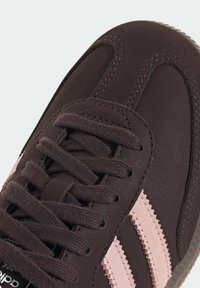 Zapatilla marrón con parte superior texturizada, franjas rosas contrastantes y cordones redondos. Suela de goma con diseño en zigzag y detalles de costura visibles.