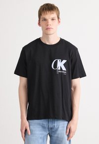 Musta puuvillapaitapaita, jossa on suuri valkoinen Calvin Klein -logo vasemmassa rintaosassa, lyhyet hihat ja rento malli.