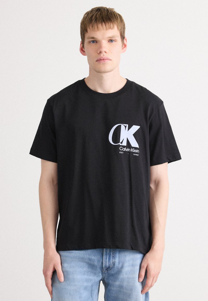 Musta puuvillapaitapaita, jossa on suuri valkoinen Calvin Klein -logo vasemmassa rintaosassa, lyhyet hihat ja rento malli.