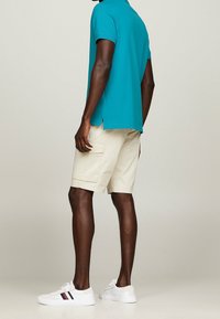 Turkusowa koszulka polo z krótkim rękawem, beżowe szorty cargo z bocznymi kieszeniami, białe sneakersy w czerwone i granatowe paski. Gładki materiał, luźny krój.