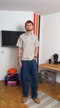 Camisa de mangas curtas, cor creme, com riscas verticais castanhas, combinada com calças de ganga azuis largas e sapatos castanhos de encaixe. Relógio simples no pulso.