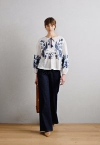 Weiße Bluse mit blauer Blumenstickerei, gekürzten Ärmeln und Bindeband am Hals, kombiniert mit dunklen Schlagjeans und geflochtenem Schuhwerk.