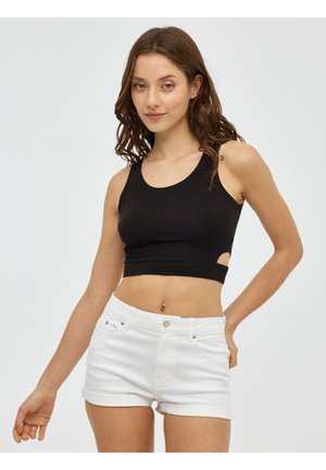 Mujer joven con cabello castaño, vistiendo un top corto negro y shorts vaqueros blancos, de pie frente a un fondo claro y liso.