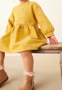 Vestido amarillo de mangas largas, con pequeños estampados de arcoíris. Tejido suave, cintura ajustada, mangas abullonadas. Botines marrones hasta el tobillo con volantes rosas.