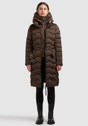 Mujer de pie con un abrigo largo acolchado marrón con un gran cuello, leggings negros y botines negros sobre un fondo gris.