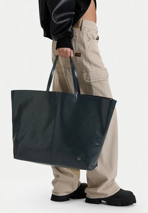 Persona con pantalones cargo beige y zapatos negros gruesos sostiene un gran bolso tote de cuero oscuro por las asas frente a un fondo liso.
