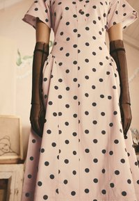 Vestido rosa con lunares y mangas cortas, con una tapeta de botones y cintura fruncida. Combinado con guantes negros transparentes para un contraste.