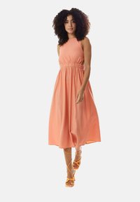 Peach-colored sleeveless dress with a fitted waist and flowy skirt. Model wears orange lace-up sandals. Smooth fabric with subtle texture.