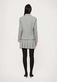 Mulher em pé de costas, vestindo um blazer cinza risca de giz e uma saia plissada, meias-calças pretas e sapatos de salto baixo pretos, contra um fundo branco.