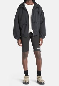 Giacca nera leggera con cappuccio, tasche frontali, polsini elastici, indossata sopra una maglietta bianca e abbinata a pantaloncini neri e sandali beige.