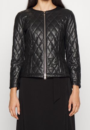 Kvinde iført en tætsiddende, sort, quiltet læderjakke med lynlås foran, kombineret med en sort nederdel og neutral baggrund.