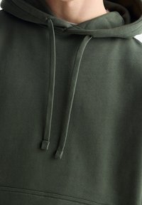 Felpa con cappuccio verde oliva con coulisse, realizzata in tessuto morbido con texture a coste e cuciture ordinate lungo l'orlo.