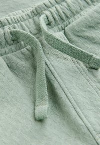 Plano detalle de unos pantalones deportivos de tela texturizada color verde claro, con cordón trenzado y cintura elástica.
