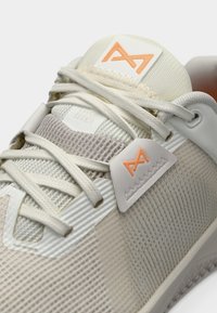 Chaussure de sport beige clair avec une tige texturée, des lacets ton sur ton, et un logo orange en accent sur la languette et le côté.