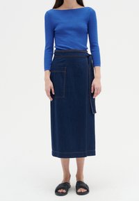 Blue textured long-sleeve top paired with a dark denim wrap skirt featuring a side pocket and contrast stitching. Black slide-on sandals.