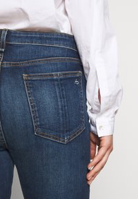 Jeans azul oscuro con un diseño clásico de cinco bolsillos, costuras visibles en un tono más claro y un pequeño logo en el bolsillo trasero.