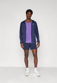 Marineblauwe lichtgewicht jas met capuchon, paarse sportshirt, marineblauwe shorts en witte schoenen met groene accenten, staand tegen een effen achtergrond.