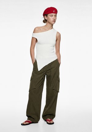 Giovane donna che indossa un top bianco asimmetrico senza maniche, pantaloni cargo color oliva, basco rosso e sandali rossi con suole nere, in piedi con le mani nelle tasche.