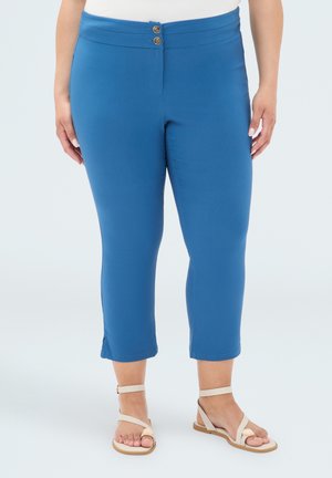 Donna che indossa pantaloni blu aderenti a gamba corta con chiusura a doppio bottone e sandali beige con cinturino alla caviglia, in piedi davanti a uno sfondo semplice.