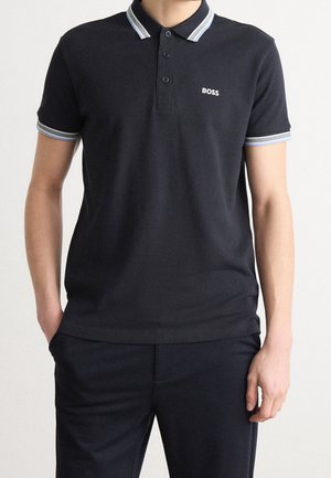 Mann trägt ein schwarzes kurzärmeliges Poloshirt mit weißem und grauem Besatz am Kragen und an den Ärmeln sowie einem "BOSS"-Logo auf der Brust.