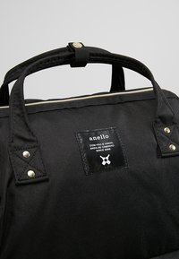 Sac à dos en toile noire avec deux poignées solides, une forme rectangulaire, des accents métalliques et un patch logo avec du texte cousu à l'avant.