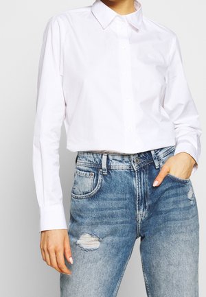 Henkilö pukeutuneena valkoiseen napilliseen paitaan, joka on tungettu vaaleansinisiin korkeavyötäröisiin kuluneisiin farkkuihin, toinen käsi taskussa.