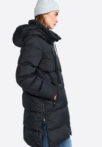 Zwarte pufferjas met een capuchon,gequilt patroon, voorrits en zijzakken. Draagt boven op blauwe jeans en vergezeld van een lichtgrijze gebreide beanie.