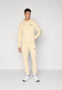 Beige hættetrøje og matchende joggers i blød stof, med et broderet logo på brystet og låret. Modellen har hvide sneakers på.