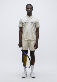 Μπεζ πόλο μπλούζα με οριζόντια γκρι ρίγα, συνδυασμένη με αντίστοιχα σορτς. Το μοντέλο κρατά ρακέτα τένις και φορά λευκές κάλτσες και αθλητικά παπούτσια.