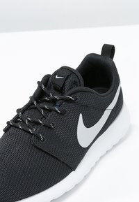 Baskets de course Nike noires avec semelle blanche et logo swoosh gris, dotées de lacets noirs avec des accents blancs sur une surface blanche.