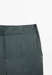 Pantalons extensibles, de couleur vert foncé, avec une taille élastique, des poches latérales et une finition en tissu texturé. Conçus pour le confort et la flexibilité.