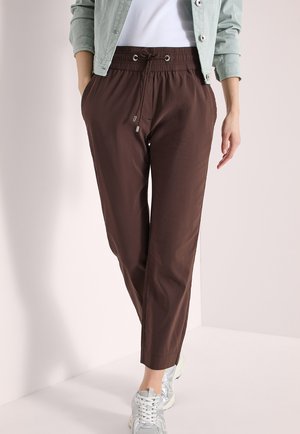 Person wearing cropped brown drawstring pants, white sneakers, white shirt, and light green buttoned jacket, standing against plain background.