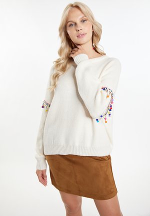 Maglione lavorato a maglia color crema con accenti di pompon colorati sulle maniche, abbinato a una mini gonna in suede marrone. Texture morbida e vestibilità rilassata.