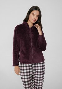 Chaqueta de forro polar burdeos con capucha, cierre completo, textura suave, combinada con pantalones de pijama de cuadros en blanco y negro.