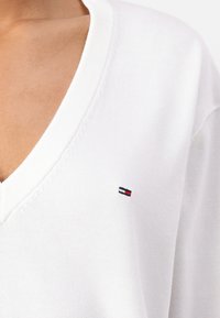 Pull blanc avec un décolleté en V, fabriqué dans un tissu lisse. Présente un petit logo brodé avec des accents rouges, blancs et bleus sur la poitrine.