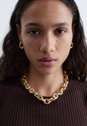 Donna con pelle media, capelli scuri e mossi, che indossa orecchini a cerchio oro, una collana a catena spessa dorata e una maglietta marrone a coste, rivolta verso l'obiettivo.