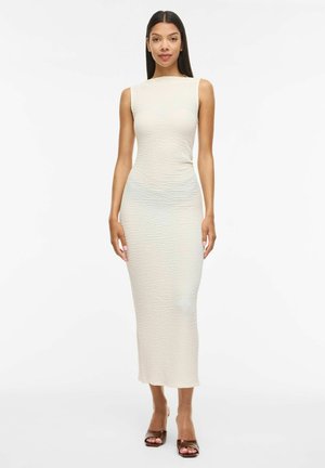 Femme aux longs cheveux foncés portant une robe midi sans manches texturée de couleur ivoire et des sandales à talons ouvertes marron, debout devant un fond blanc.