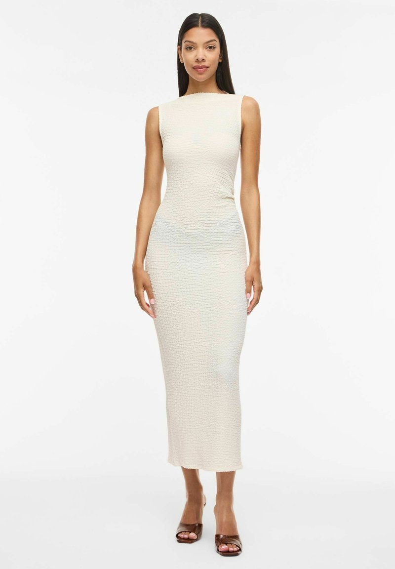 Femme aux longs cheveux foncés portant une robe midi sans manches texturée de couleur ivoire et des sandales à talons ouvertes marron, debout devant un fond blanc.