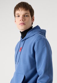 Blå hettegenser med frontlomme, rød logo-lapp og snøring. Stoffet ser mykt ut med en jevn tekstur.