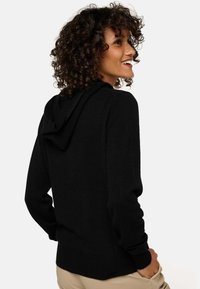 Femme aux cheveux bouclés portant un sweat à capuche noir et un pantalon beige, souriante et regardant vers la droite sur fond blanc.