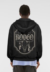 Černá mikina s kapucí s velkým grafickým designem na zadní straně, zobrazující lebku a peří se slovem "RODEO" ve stylizovaném písmu.