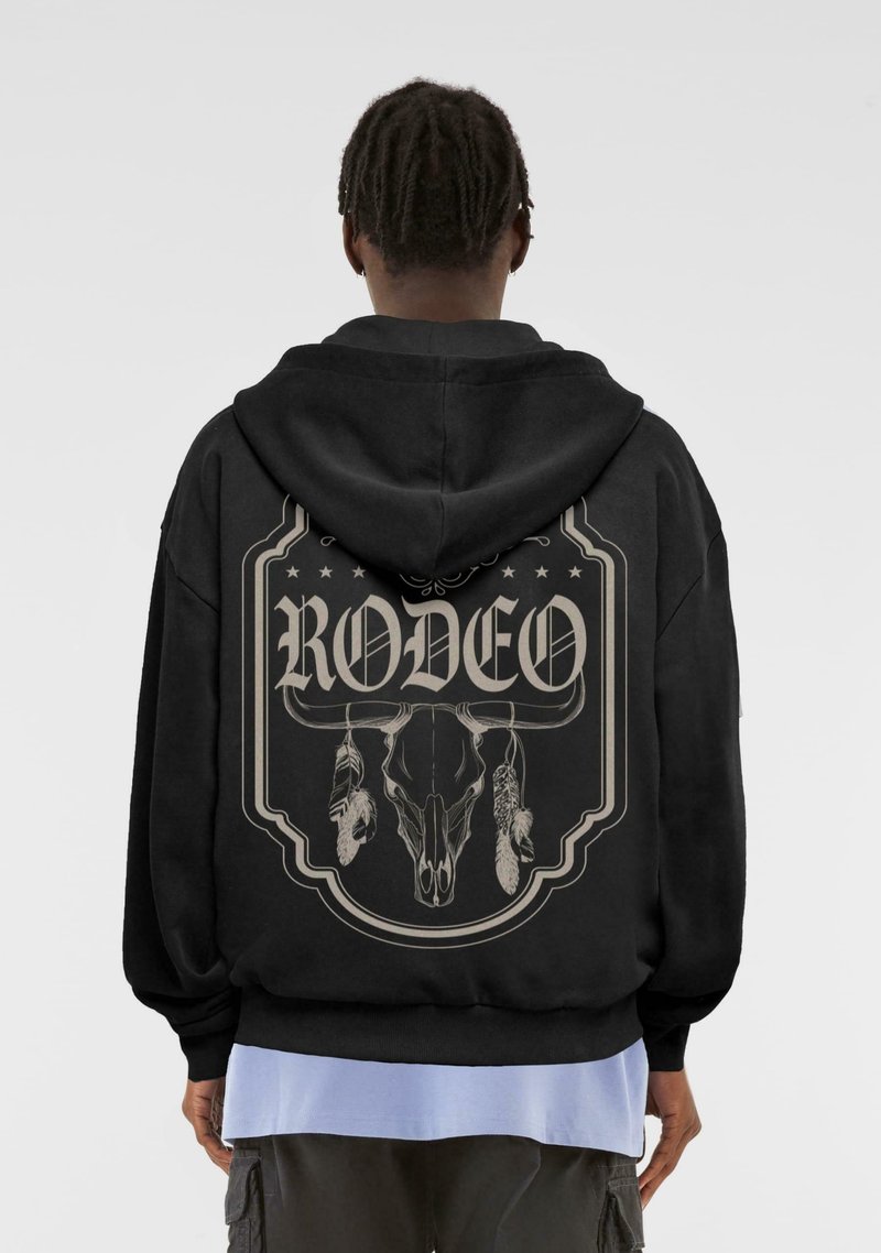 Černá mikina s kapucí s velkým grafickým designem na zadní straně, zobrazující lebku a peří se slovem "RODEO" ve stylizovaném písmu.