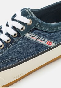 Tenis de denim com um cabedal azul escuro, atacadores brancos e uma sola de borracha com uma sola de goma. Inclui uma etiqueta de logotipo da marca na lateral.