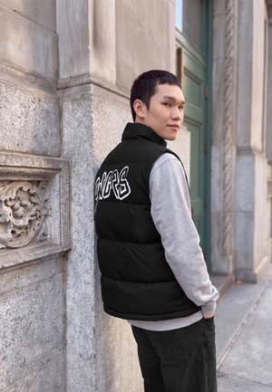 Jeune homme portant un gilet matelassé noir avec un texte blanc, un sweat-shirt gris et un pantalon noir, debout contre un mur en pierre détaillé et regardant en arrière.