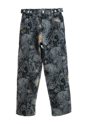 Joven con chaqueta y pantalones a juego con un patrón de noche estrellada en remolino y botas marrones, de pie sobre fondo blanco.