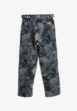 Joven con chaqueta y pantalones a juego con un patrón de noche estrellada en remolino y botas marrones, de pie sobre fondo blanco.