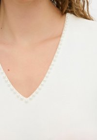 Top blanco con escote en V adornado con pequeños acentos de perlas falsas, espaciados uniformemente alrededor del cuello. Textura de tela suave.