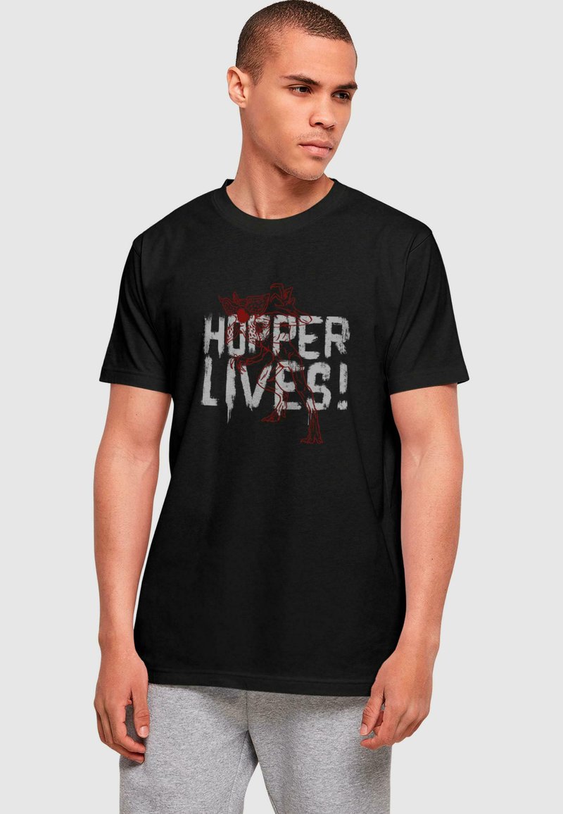 T-shirt en coton noir avec un imprimé graphique audacieux, "Hopper Lives !" en blanc et rouge, accompagné d'une illustration de personnage. Manches courtes, col rond.