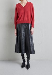 Kvinna iklädd en röd V-ringad tröja, svart läder midi-kjol och svarta spetsiga stövlar med klack, stående mot en enkel ljusgrå vägg.